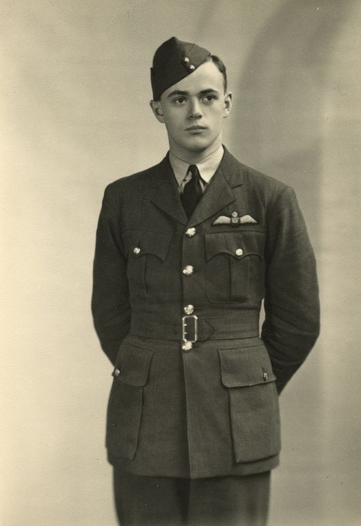 Studio portrait of a young man in Royal Air Force uniform, standing with hands behind his back, wearing a side cap and displaying pilot wings on his chest