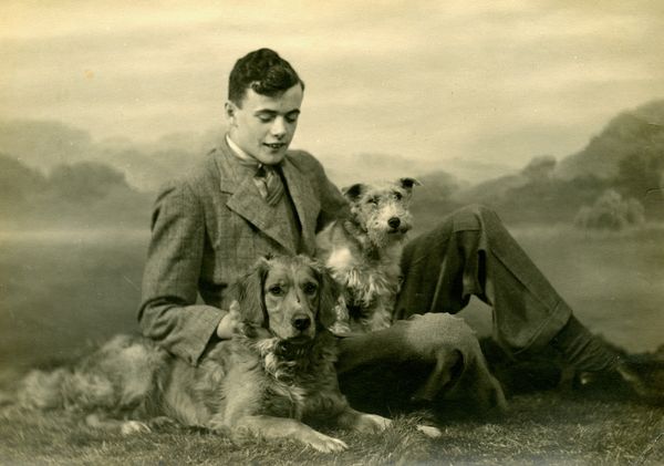 Vintage photograph of a young man in a suit sitting on grass with two dogs, one shaggy terrier on his lap and a spaniel lying in front, against a painted countryside backdrop